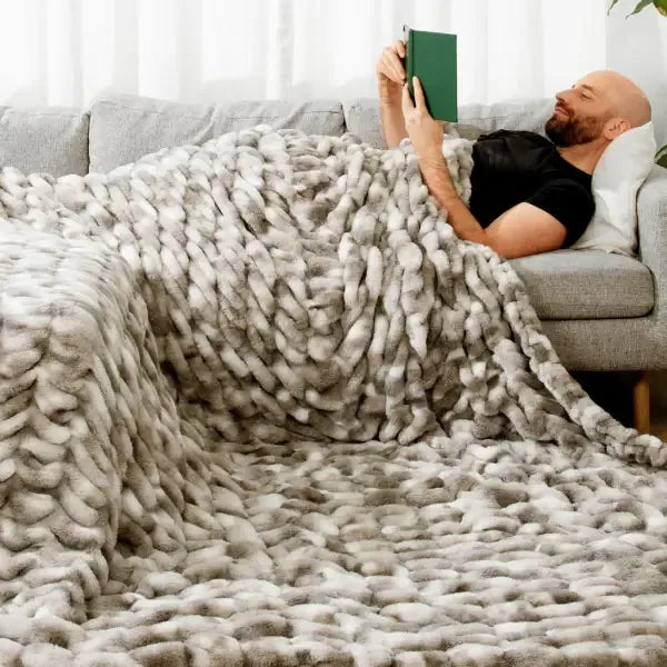 Man relaxed on gray sofa under chunky knit cream oversized blanket while reading