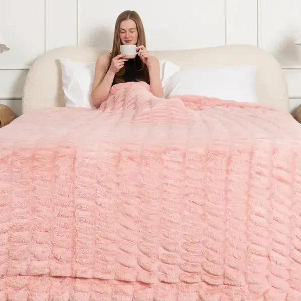 Woman sitting on bed holding phone under oversized rose pink textured blanket with quilted pattern
