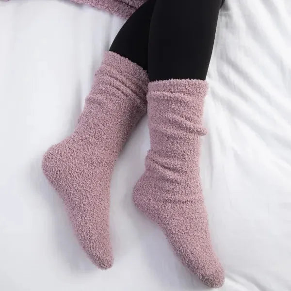Person wearing mauve fuzzy socks on white blanket, black pants visible