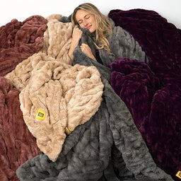 Woman wrapped in oversized Cloud Luxe blanket in charcoal gray surrounded by display of blankets in burgundy, cream, and plum colors