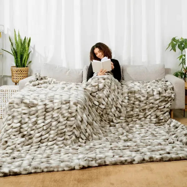 Woman reading under oversized chunky knit cream blanket on couch with plants