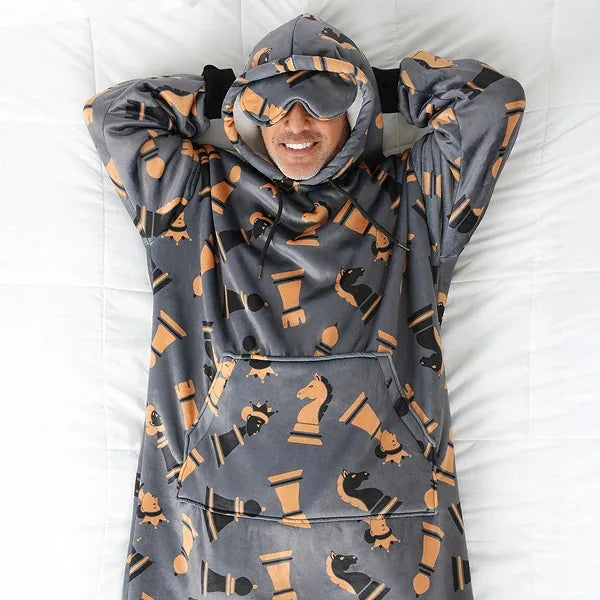 Person wrapped in gray and orange chess-patterned oversized hoodie blanket with hood and front pocket, smiling overhead view on white bedding