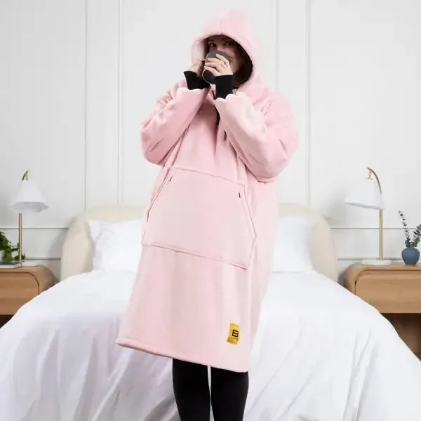 Person wearing pink oversized Hideout Hoodie blanket with hood up, standing on white bedding in modern bedroom