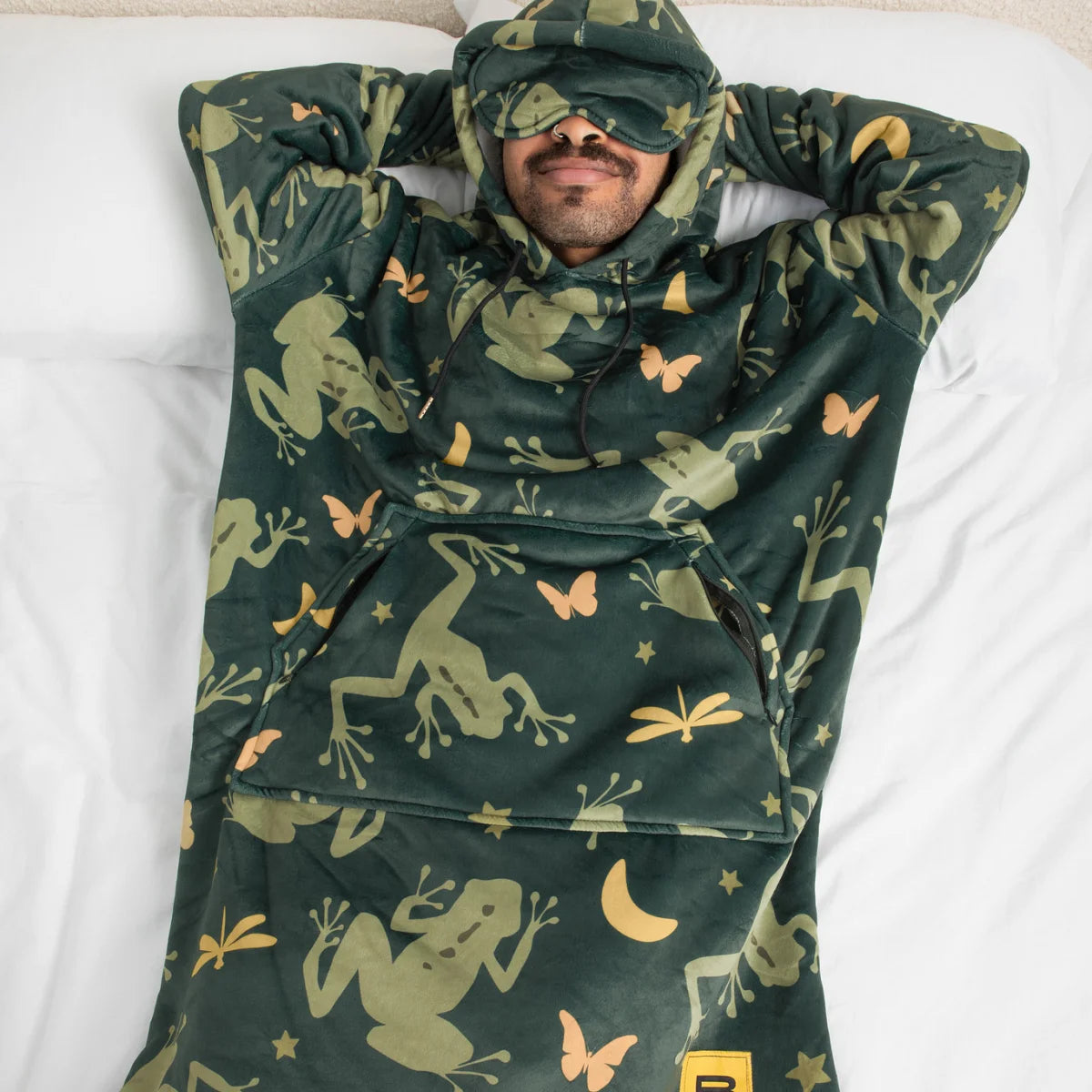 Person lying in bed in the Dreamy Frogs Hideout Hoodie™ with the built-in eye mask pulled down
