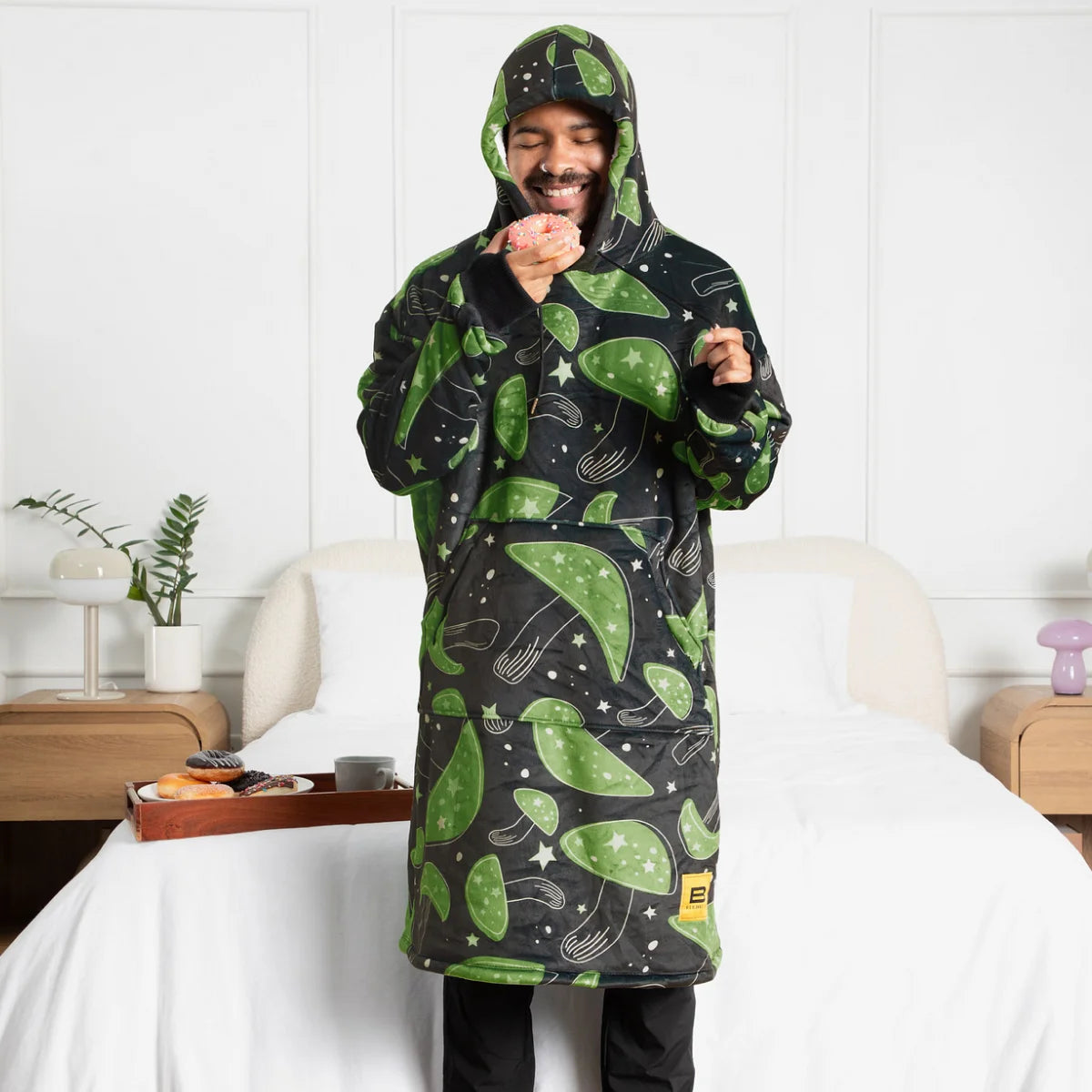 Person standing in a bedroom wearing the Mystic Mushrooms Hideout Hoodie™ with the hood up, holding a donut