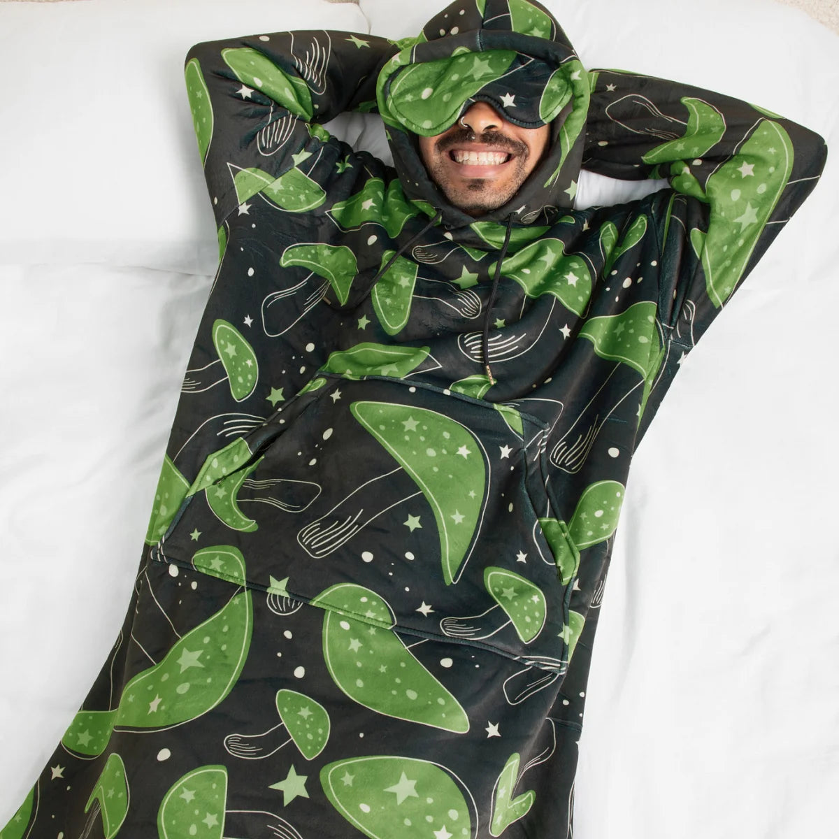 Person lying back in bed in the Mystic Mushrooms Hideout Hoodie™ with the built-in eye mask pulled down and arms behind their head