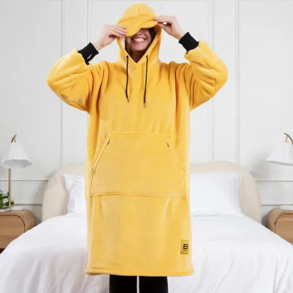 Person wearing yellow oversized Hideout Hoodie with hood and matching cap, standing on bed with white pillows