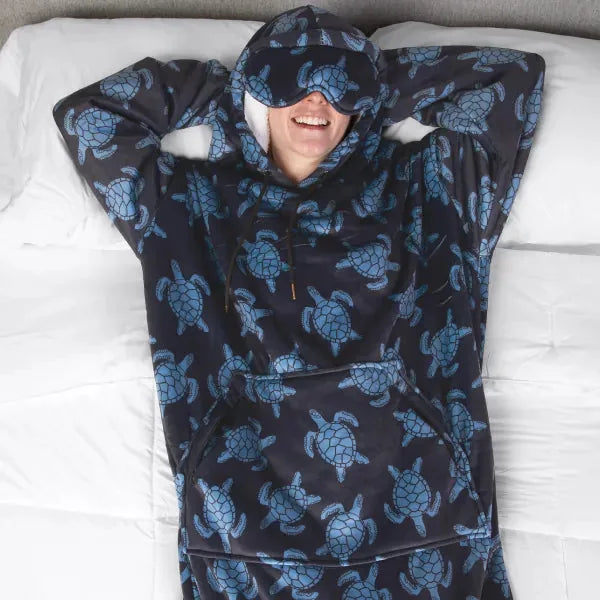 Person wrapped in navy oversized blanket with blue turtle print, wearing matching hood and eye mask, lying on white bedding