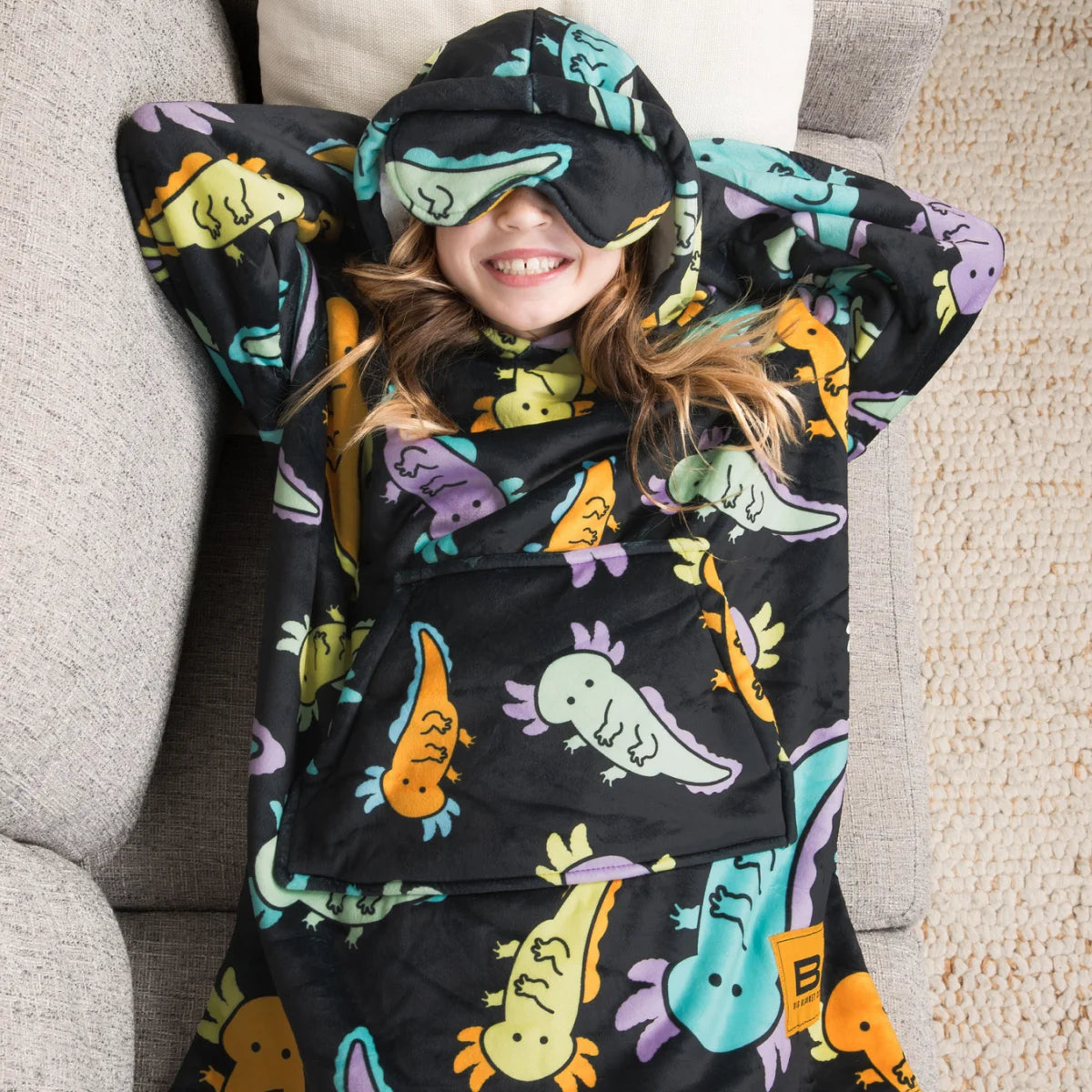 Child relaxing on a couch in the Axolotl Drift Hideout Hoodie™ Kids wearable blanket with the eye mask down