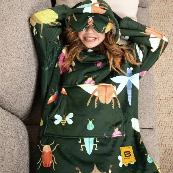 Child wrapped in forest-green oversized blanket hoodie with colorful bug and insect print, wearing matching eye mask