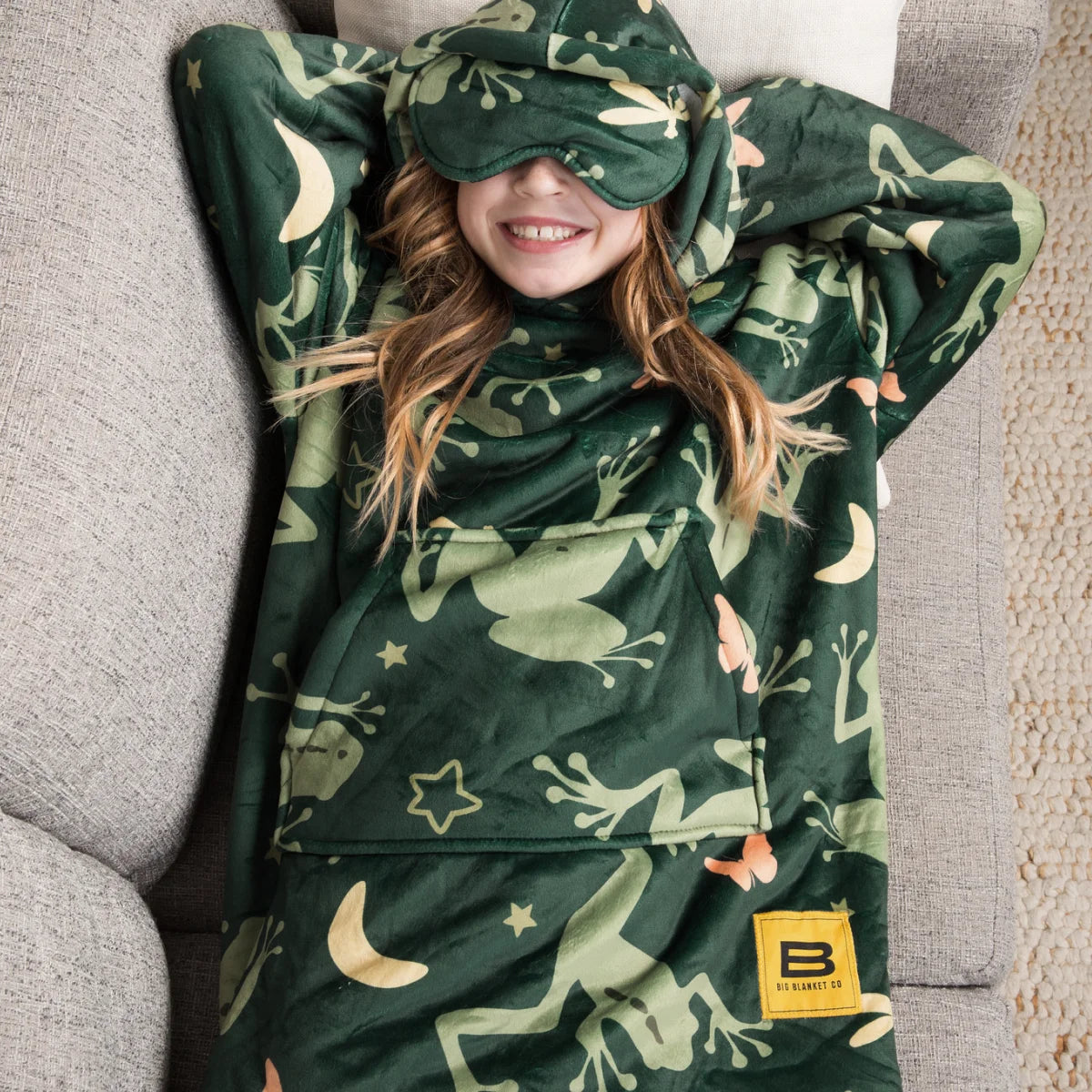 Child relaxing on a couch in the Dreamy Frogs Hideout Hoodie™ Kids wearable blanket with the eye mask down