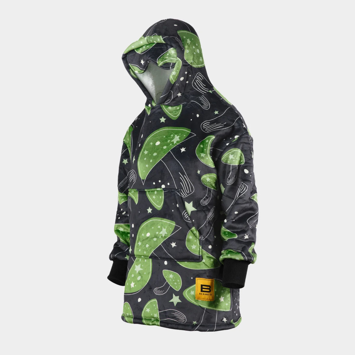 Hideout Hoodie™ Kids wearable blanket in Mystic Mushrooms, side view with the hood up and the built-in eye mask on the hood — near-black charcoal ground scattered with large green mushroom caps, cream stars and speckles