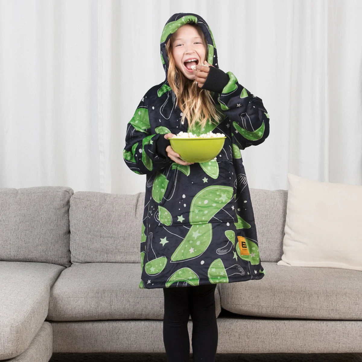 Child standing in front of a couch in the Mystic Mushrooms Hideout Hoodie™ Kids with the hood up, holding a bowl of popcorn