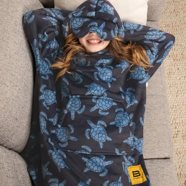 Child wrapped in oversized navy blanket with blue turtle print, peeking over fabric with hands behind head on couch