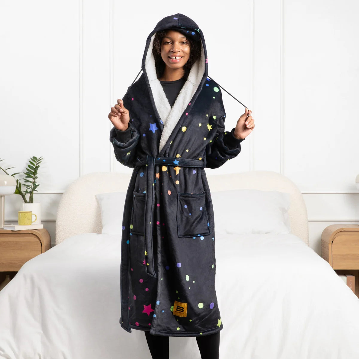 Person standing beside a bed in the Electric Sky Hideout Robe™ with the sherpa-lined hood up