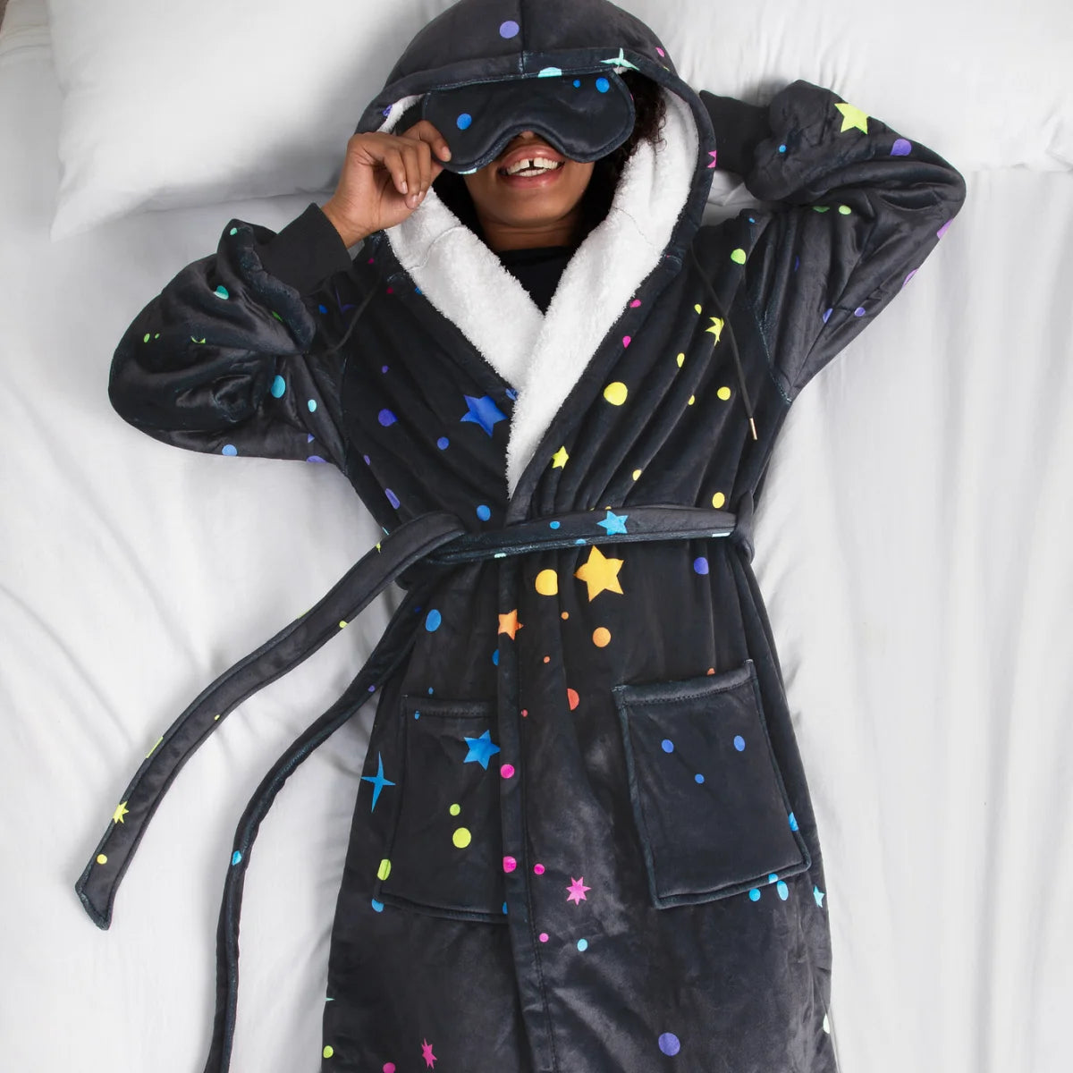 Person lying on white bedding in the Electric Sky Hideout Robe™ with the built-in eye mask on
