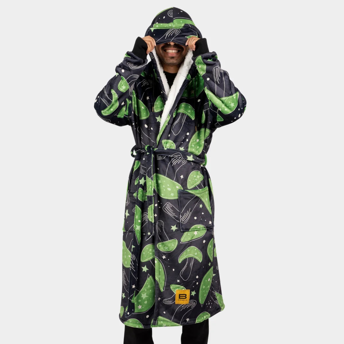 Person facing the camera lifting the built-in eye mask of the Mystic Mushrooms Hideout Robe™, sherpa-lined hood and lapel visible