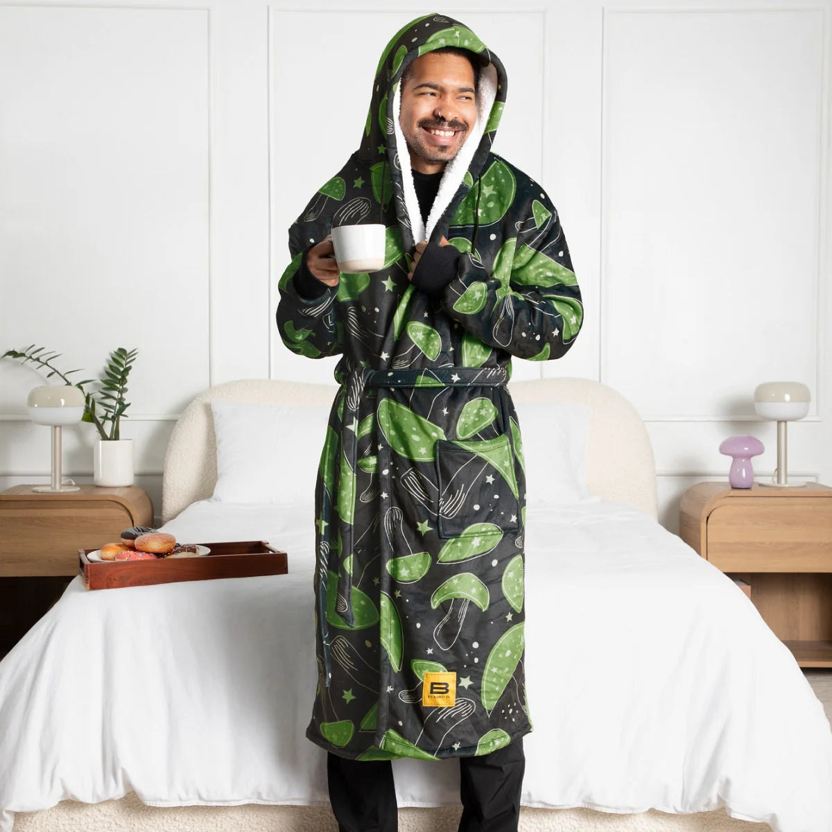 Person standing beside a bed in the Mystic Mushrooms Hideout Robe™ with the sherpa-lined hood up, holding a mug