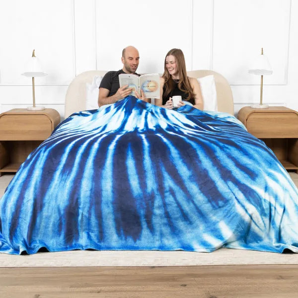 Couple sitting on bed under large blue tie-dye oversized blanket, reading together in modern bedroom