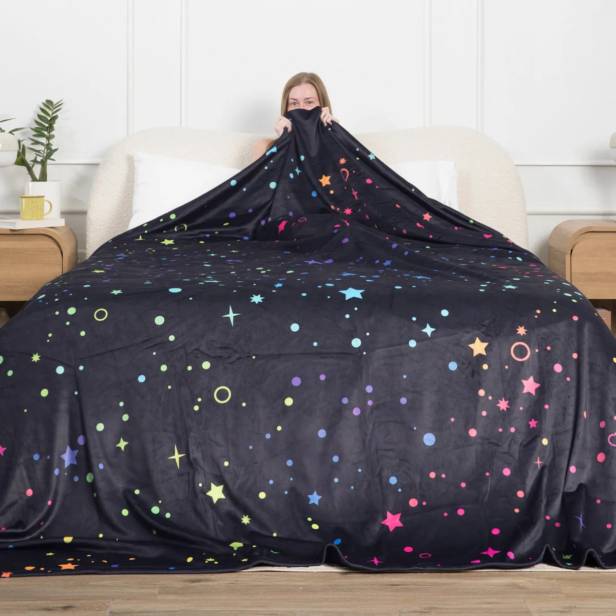 Person sitting up in bed holding the Electric Sky Original Stretch™ oversized blanket up over a made bed