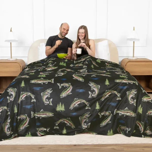 Couple sitting on bed under dark fishing-themed oversized blanket with fish and forest patterns, holding mugs