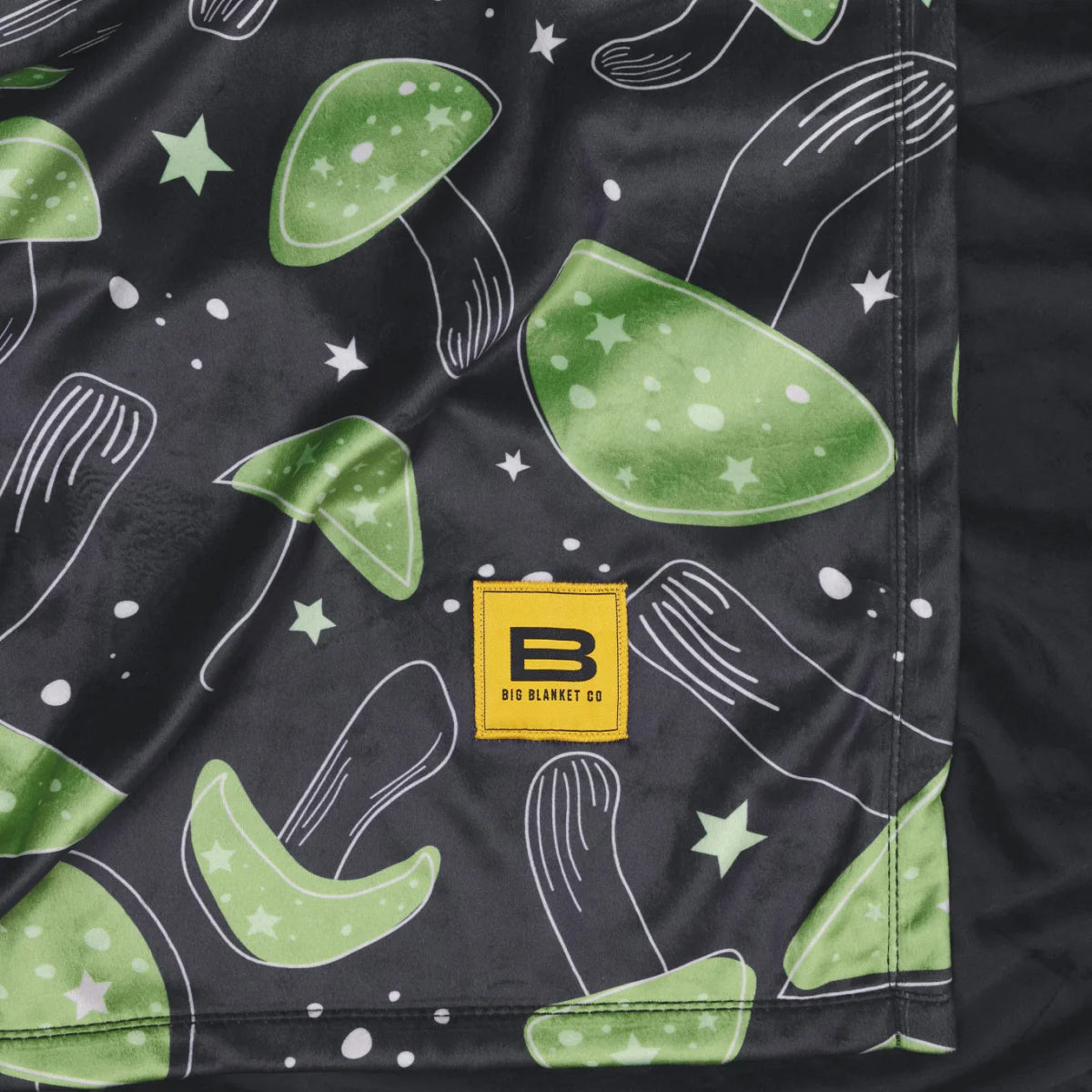 Close-up of a corner of the Mystic Mushrooms Original Stretch™ blanket showing the green mushroom print and the yellow Big Blanket Co label