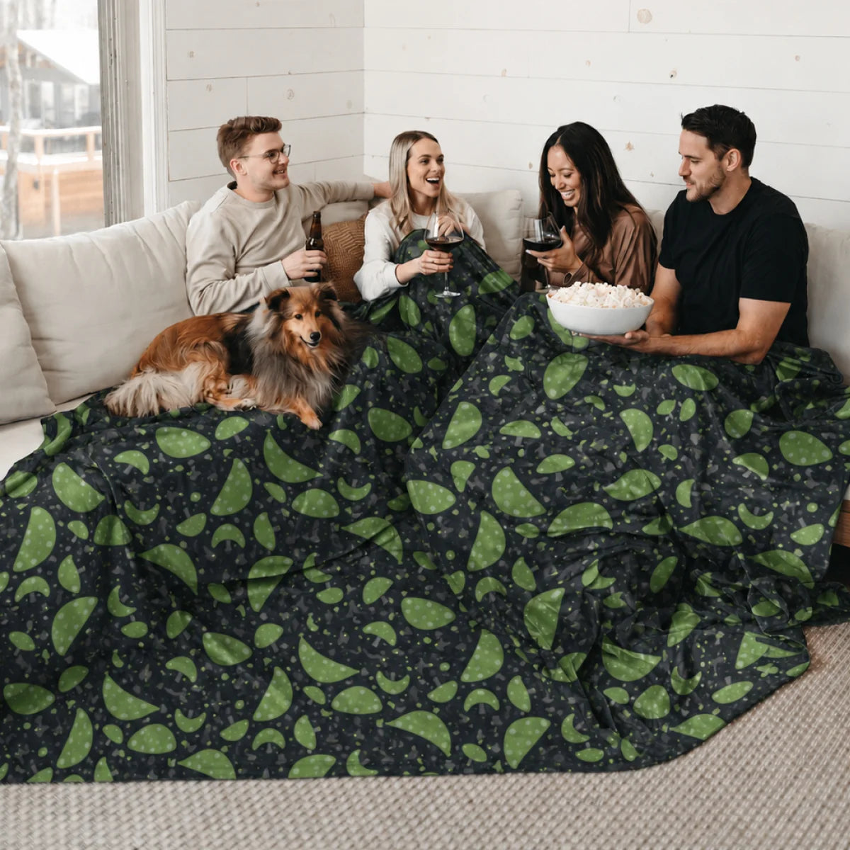 Four friends and a dog sharing two Mystic Mushrooms Original Stretch™ oversized blankets on a couch with popcorn and wine