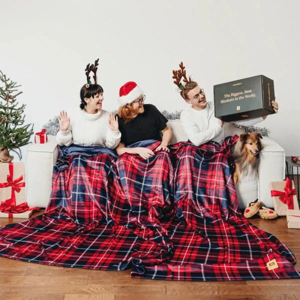 Three people and dog snuggled under red plaid oversized blanket with holiday decorations and gifts nearby