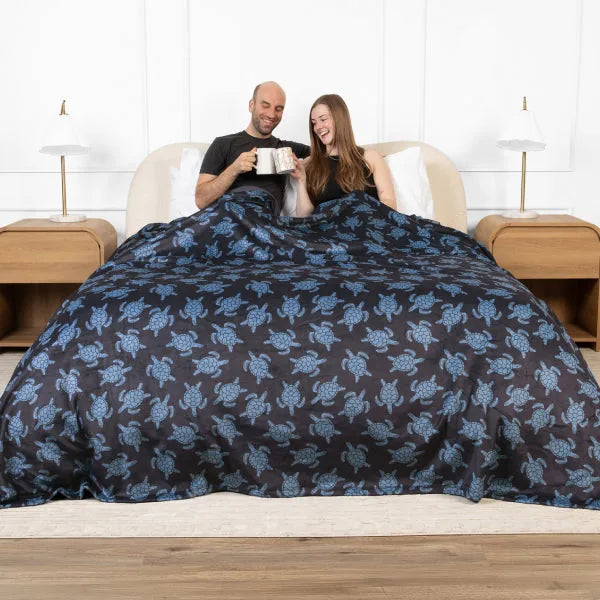 Couple in bed under navy oversized blanket with blue floral pattern, holding mugs together
