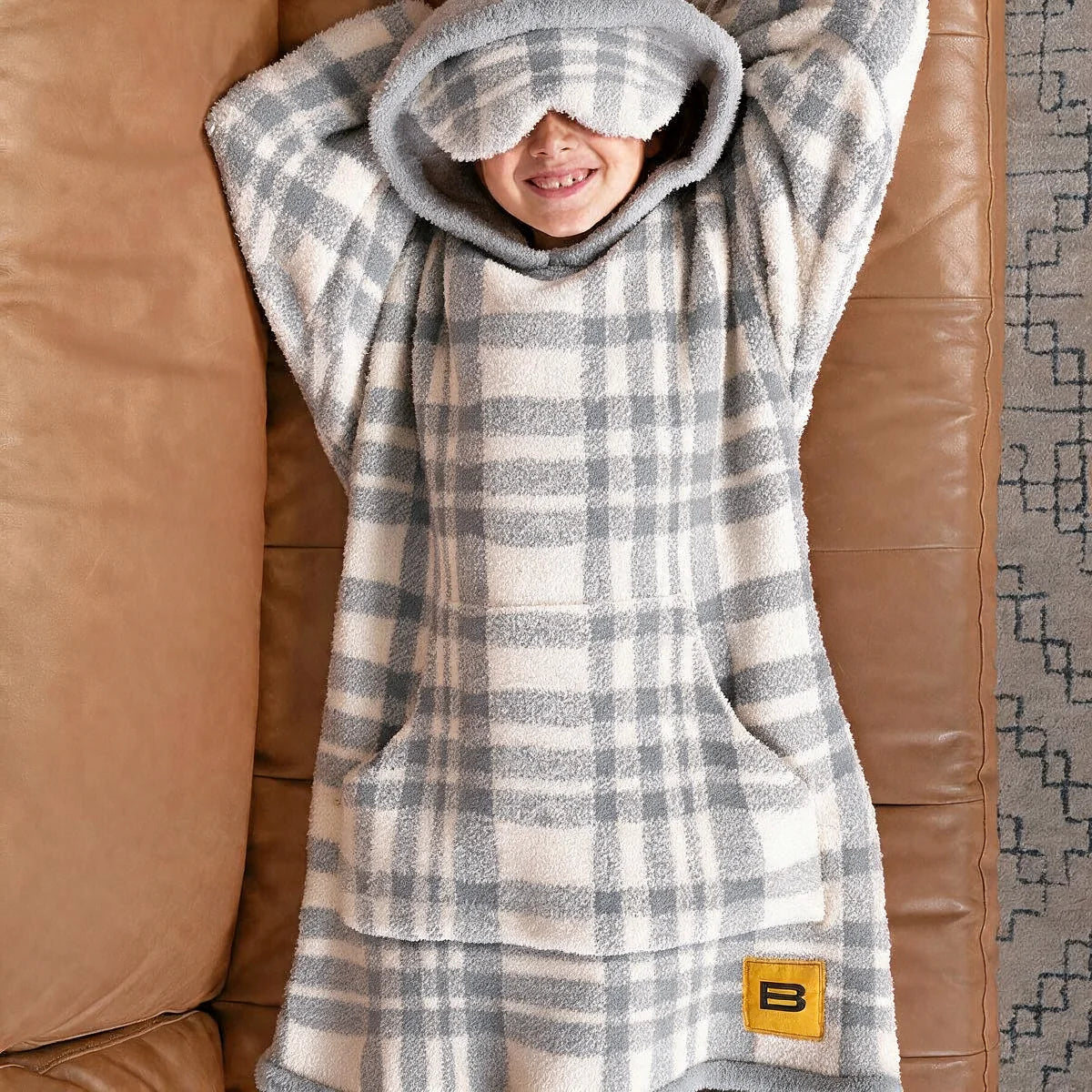 Child standing in front of a couch wearing the Grey Plaid Premier Plush™ Hoodie Kids with the hood up