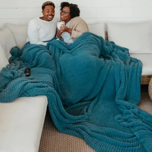 Two people bundled under oversized teal quilted blanket on white sofa, holding hot drinks