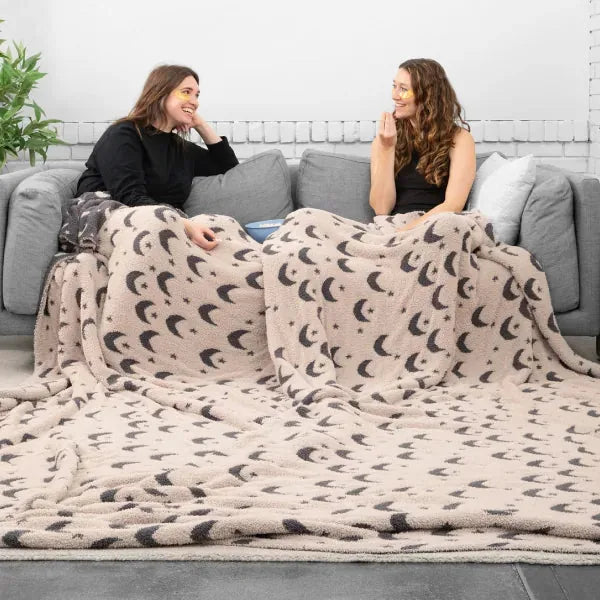 Two women sitting on gray couch wrapped in cream oversized blanket with moon pattern, demonstrating size and comfort