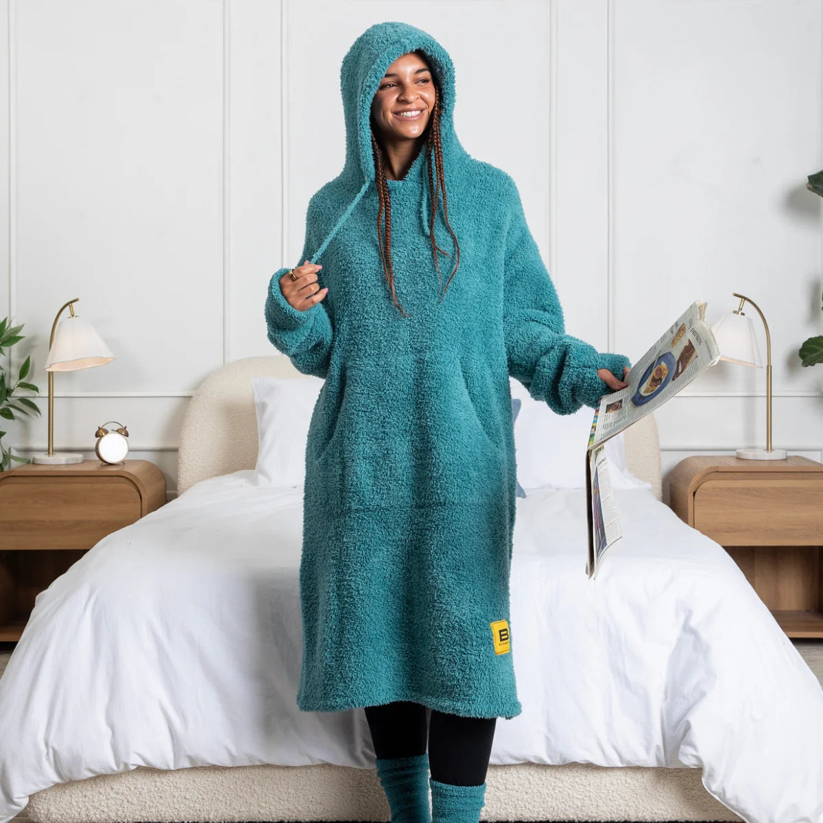 Woman wearing teal oversized hoodie blanket with hood and kangaroo pocket, holding phone on couch
