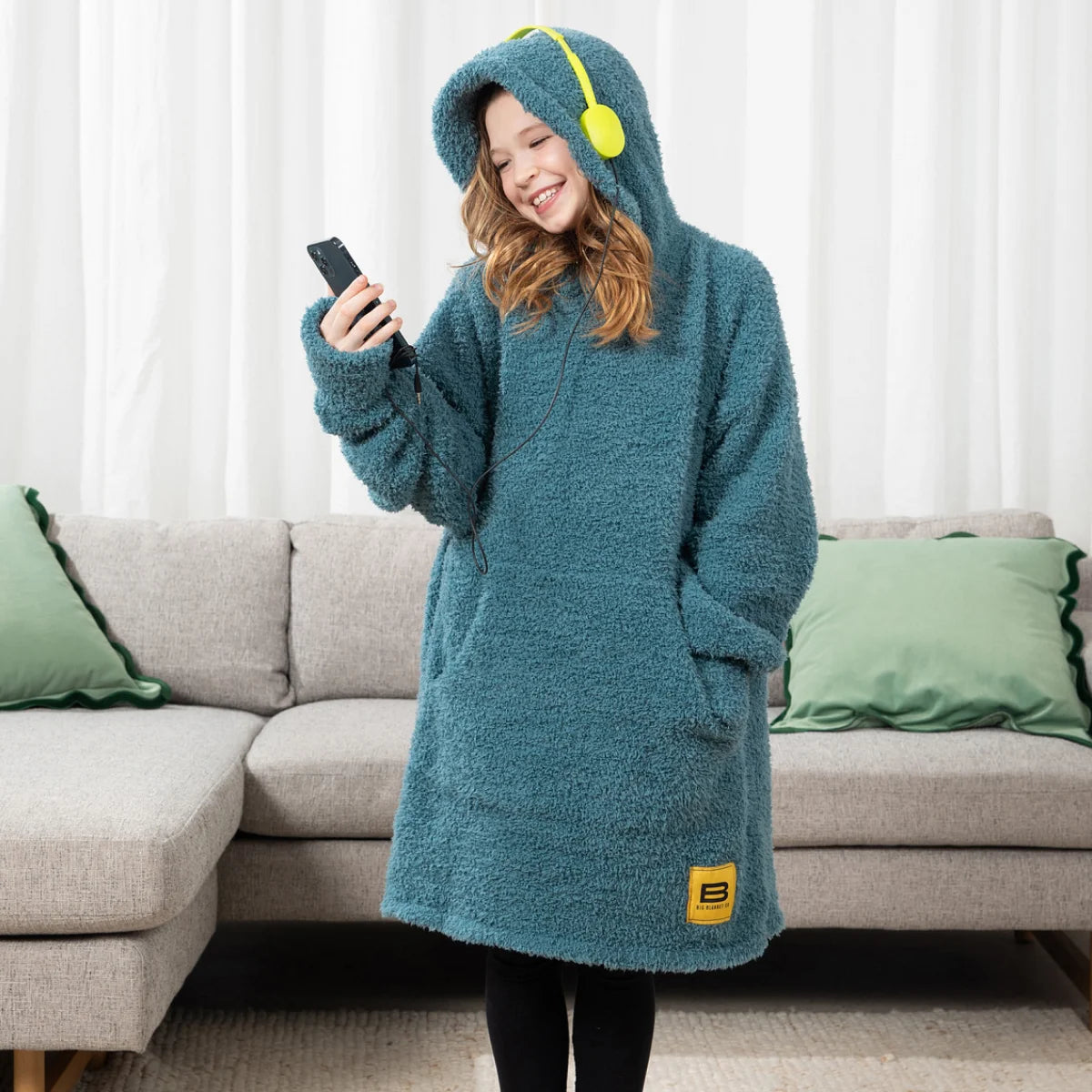 Child wearing teal Premier Plush Hoodie with hood up, holding phone and wearing headphones, standing in front of couch