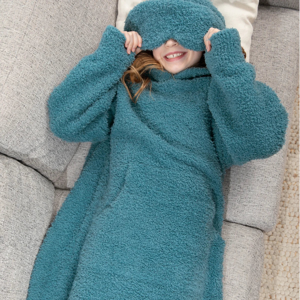 Child wrapped in teal oversized hoodie blanket on gray couch, covering eyes with hood