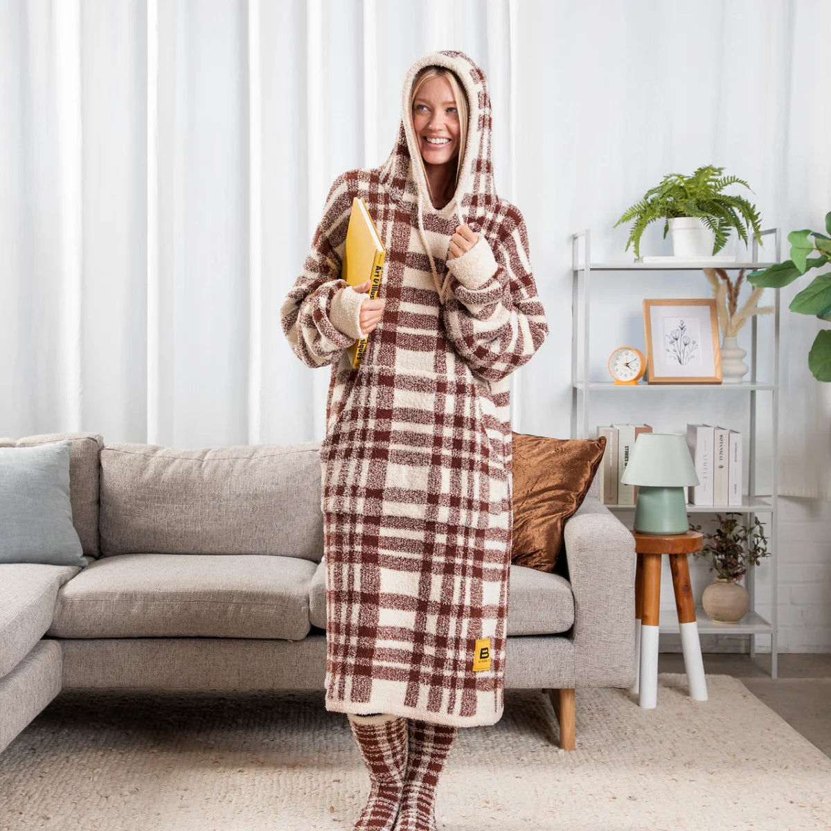 Woman wearing burgundy plaid oversized hoodie blanket with cream stripes, standing in living room with couch
