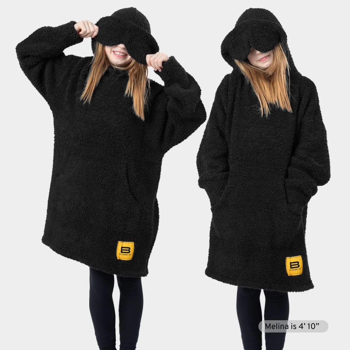 Black oversized hoodie blanket with kangaroo pocket and drawstring hood, shown on model in two poses with hands in pockets