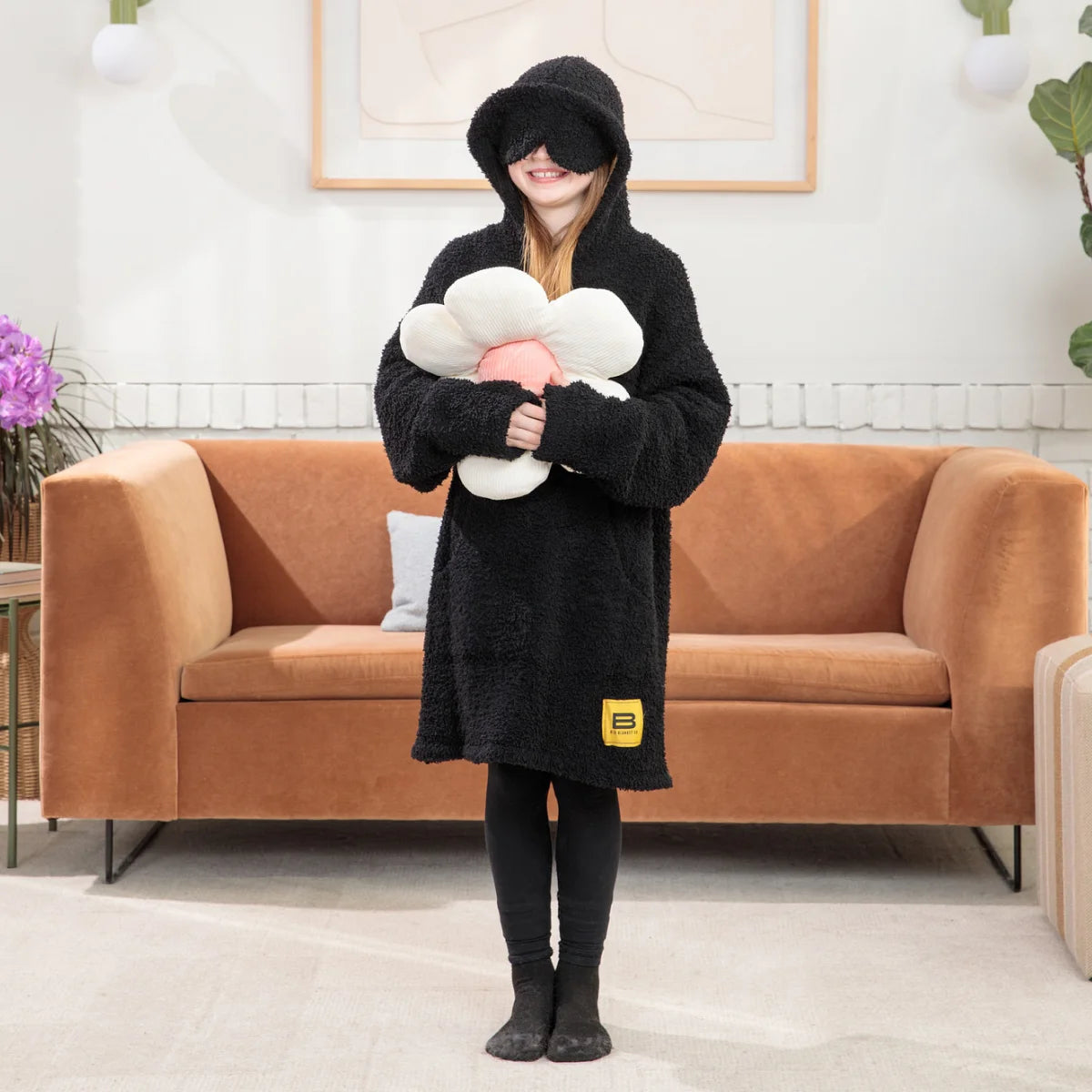 Woman wearing black oversized hoodie blanket with hood up, holding bowl and spoon, standing in front of bed