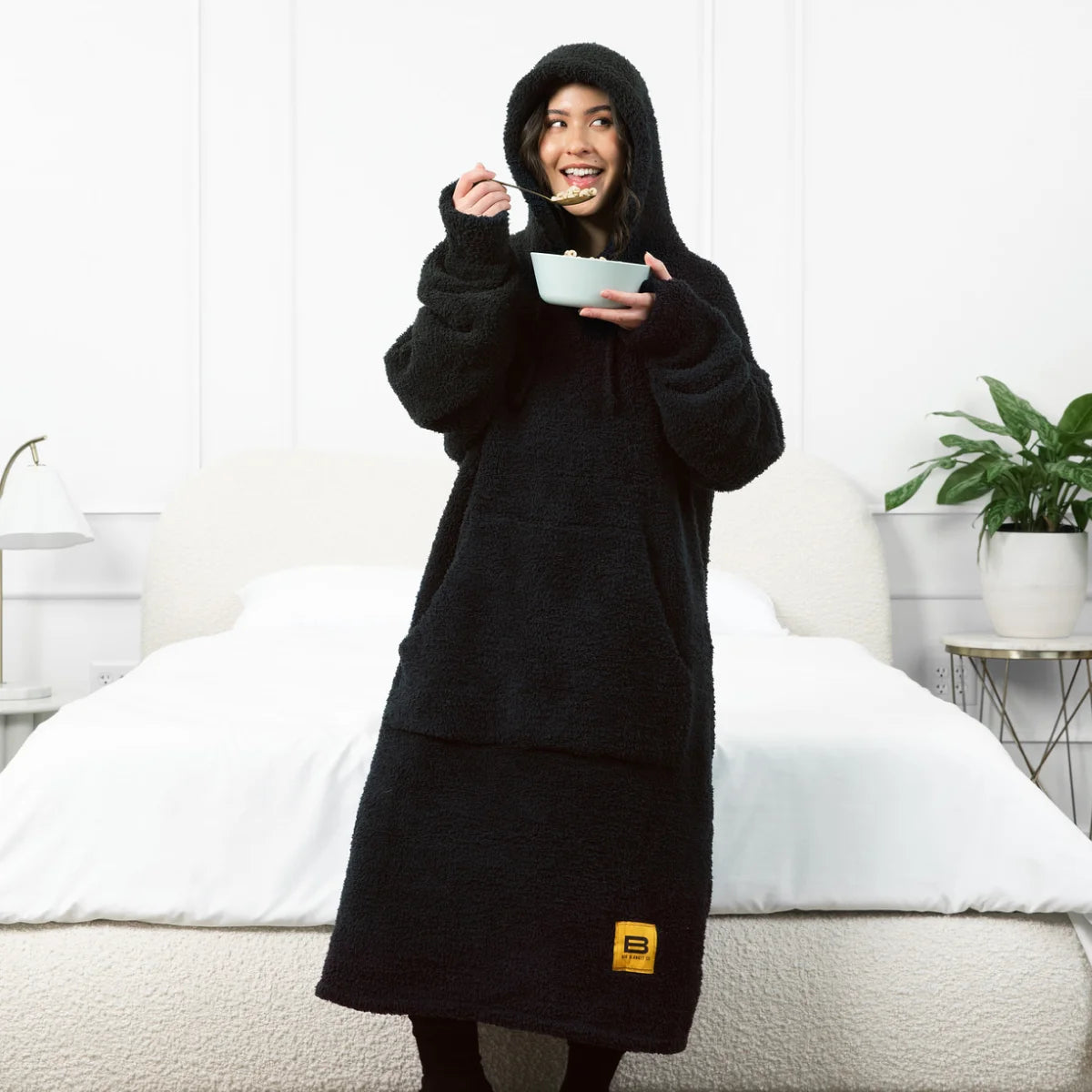 Woman wearing black Premier Plush oversized hoodie blanket with attached hood, eating from bowl while standing in bedroom