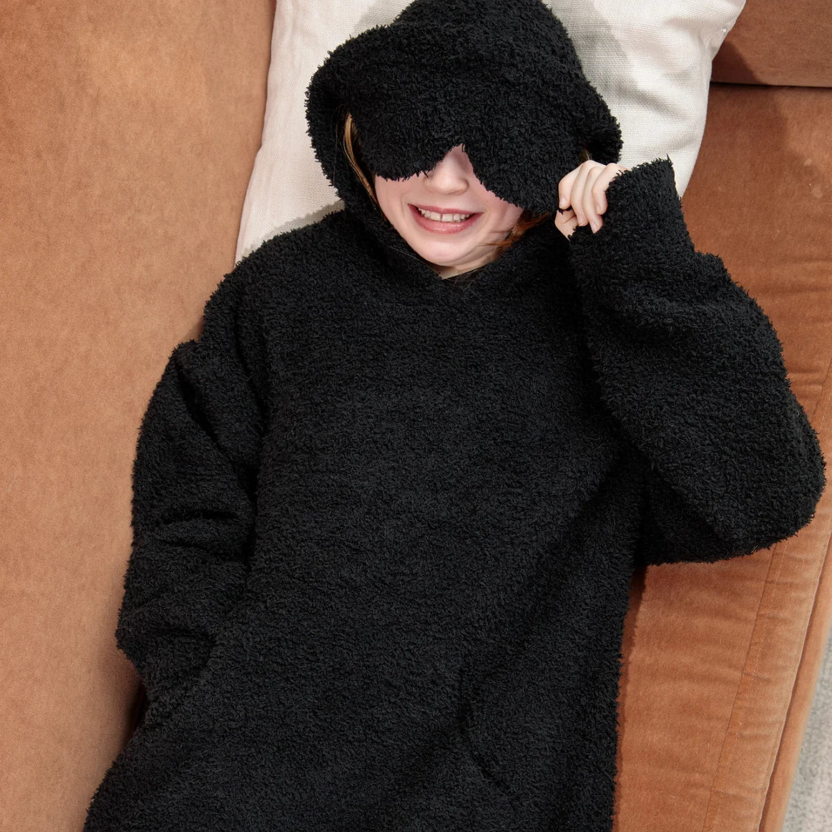 Man wrapped in oversized black hoodie blanket with white bedding, hood covering eyes playfully
