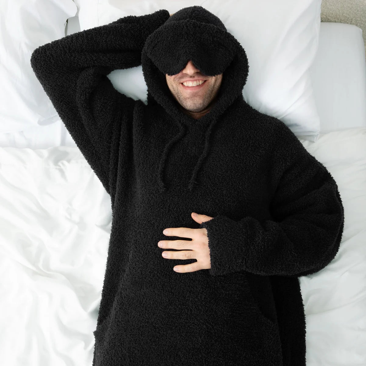 Man wrapped in black oversized hoodie blanket with hood covering eyes, relaxing on white bedding