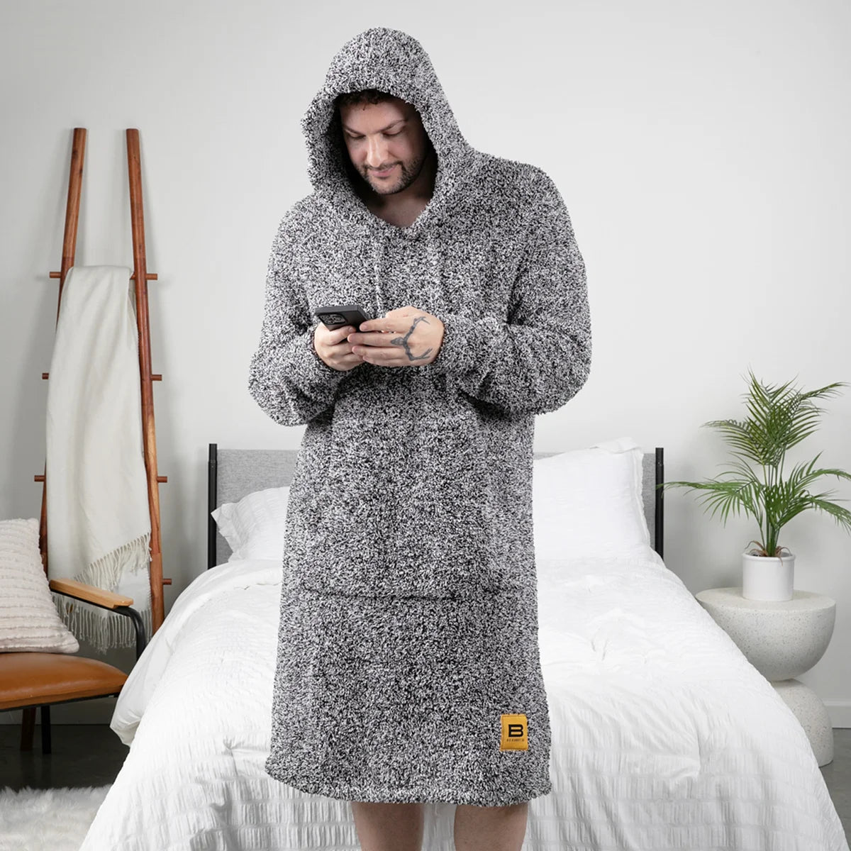 Man wearing charcoal heather oversized hoodie blanket with hood up, standing on bed holding phone
