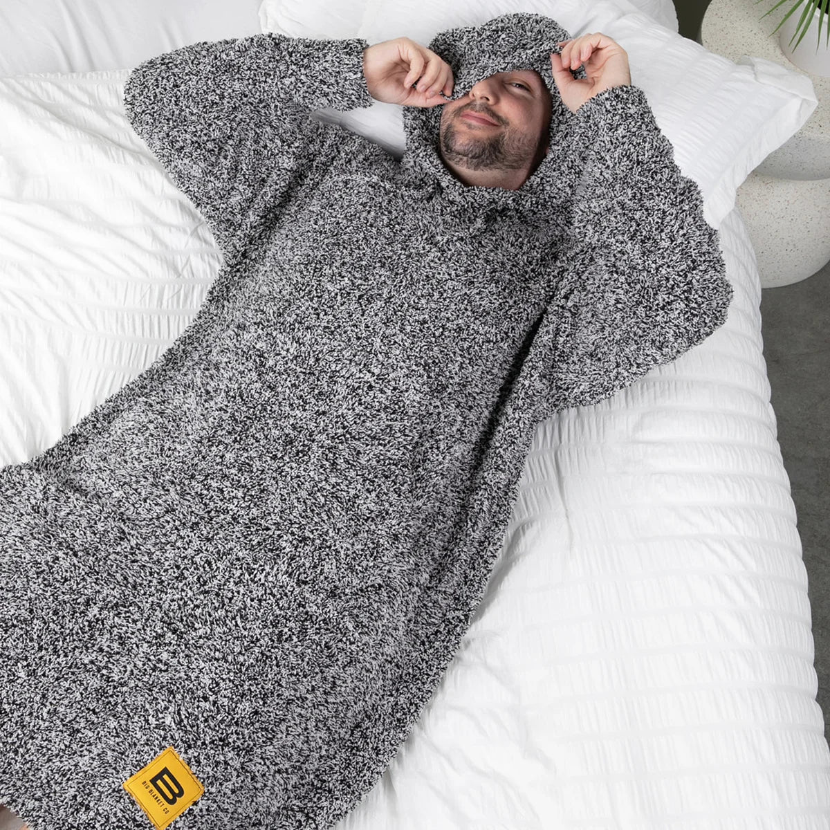 Man wrapped in charcoal heathered oversized hoodie blanket on white bedding, overhead view showing plush texture and coverage