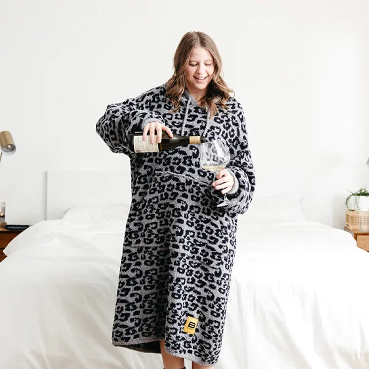 Woman wearing black leopard print oversized hoodie blanket, pouring wine while standing on bed
