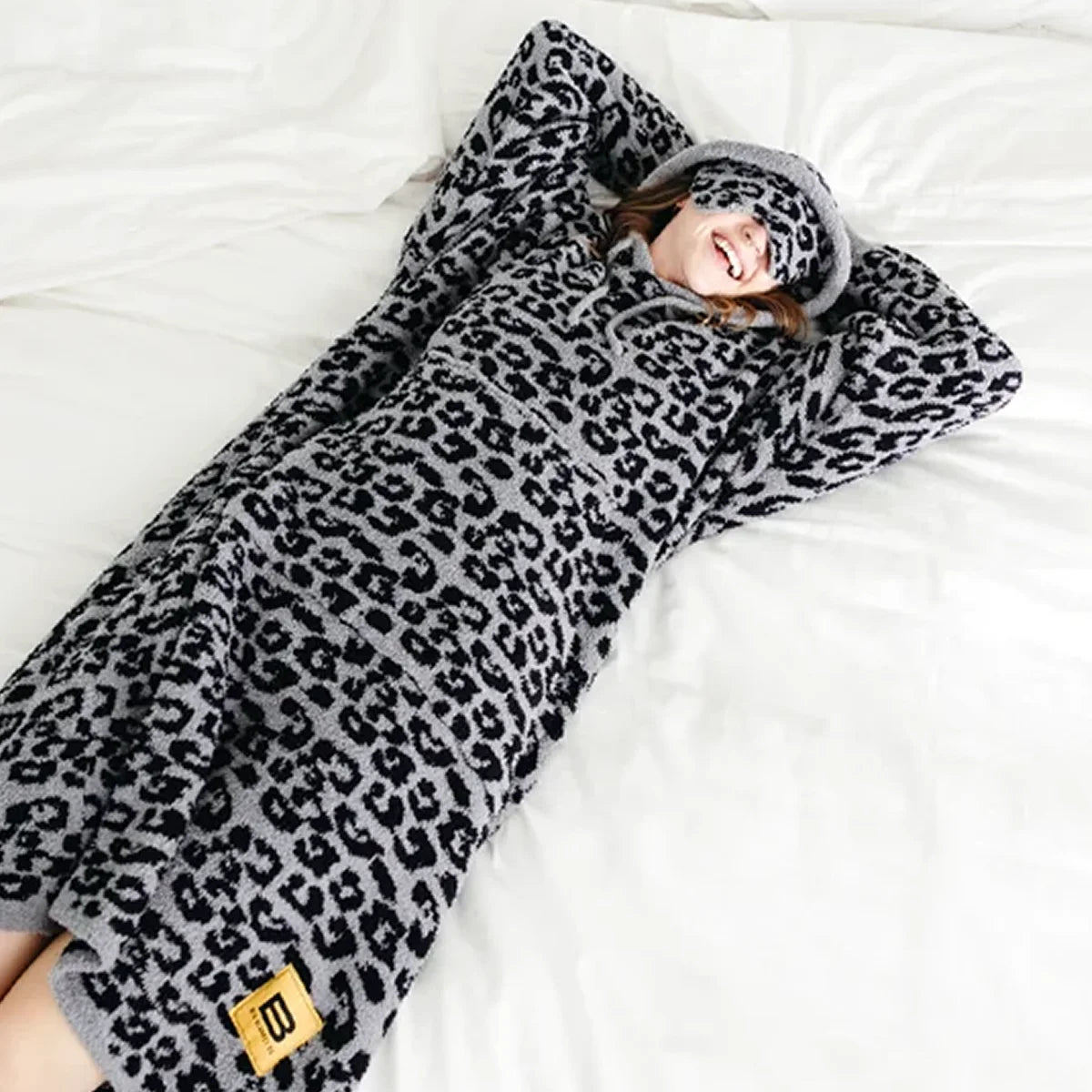 Woman wrapped in black leopard print oversized hoodie blanket on white bedding, smiling