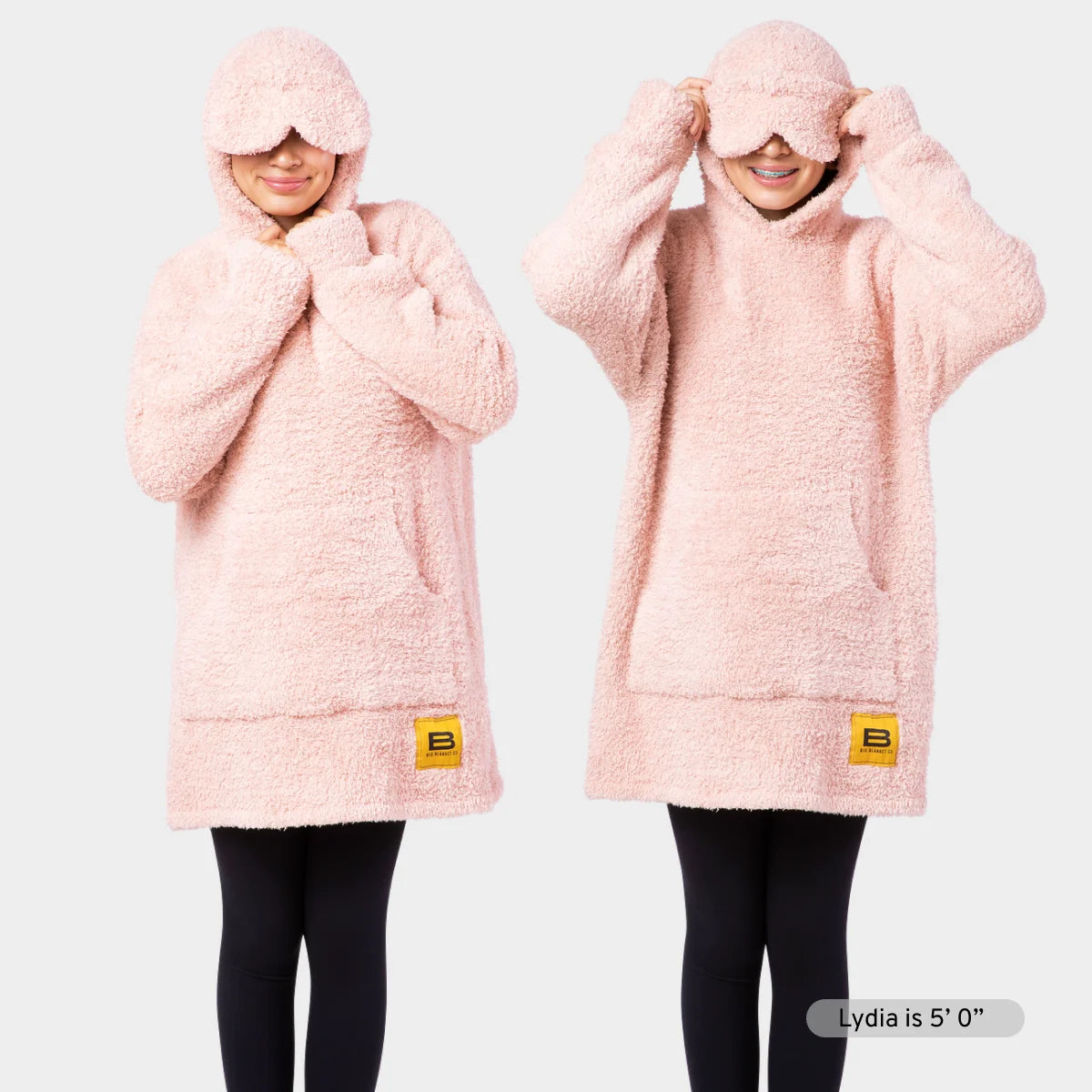Pink oversized hoodie blanket with kangaroo pocket and drawstring hood, shown on two models wearing matching socks