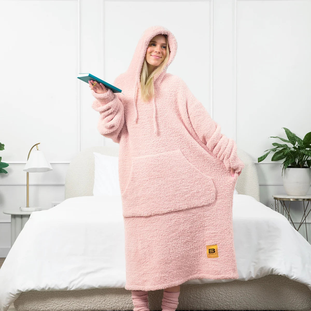 Woman wearing blush pink oversized hoodie blanket with kangaroo pocket, reading on bed