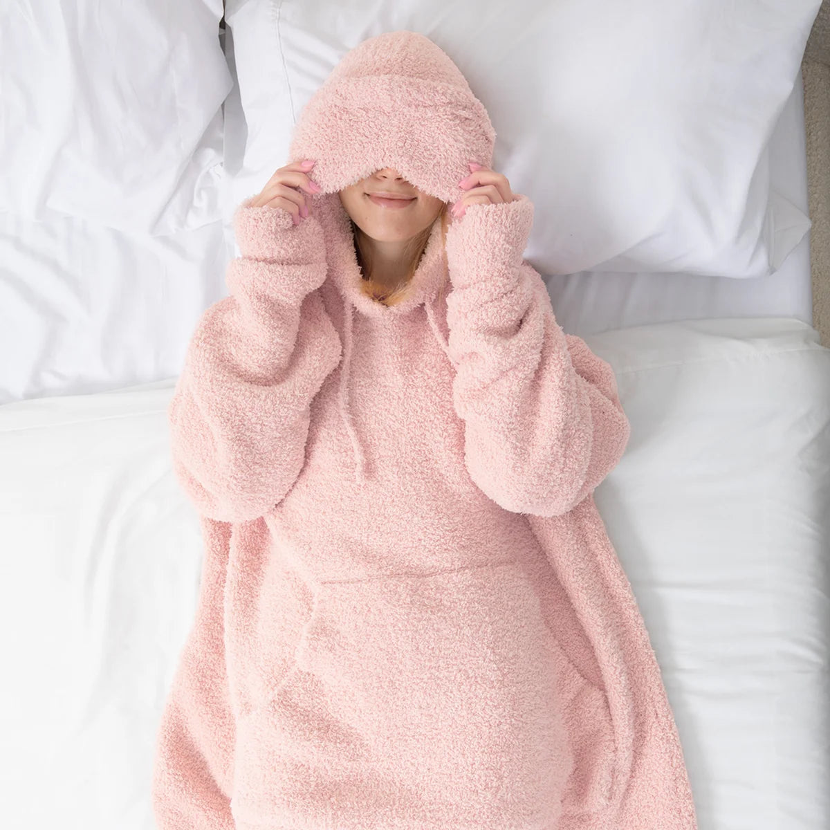 Child wrapped in blush pink oversized hoodie blanket on white bedding, hood covering eyes