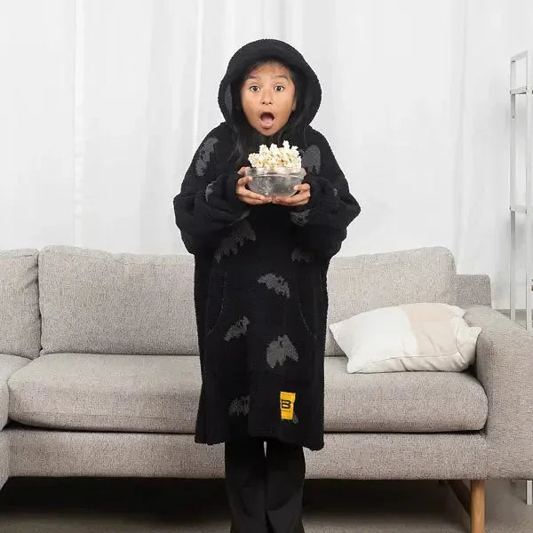 Child wearing black oversized blanket hoodie with bat pattern, holding popcorn bowl, standing in front of gray couch