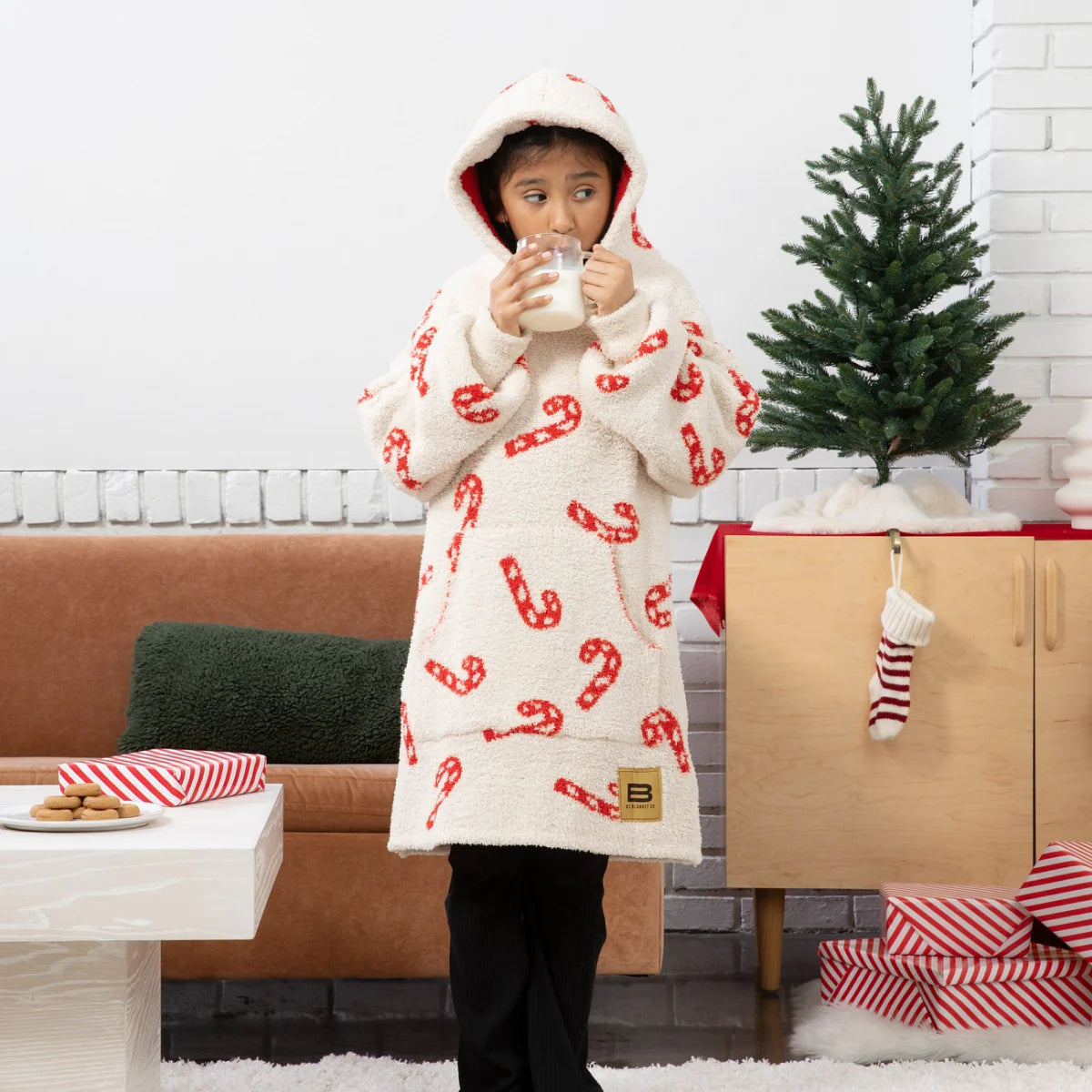 Woman wearing cream oversized hoodie blanket with red candy cane pattern, modeling with matching hood up on bed